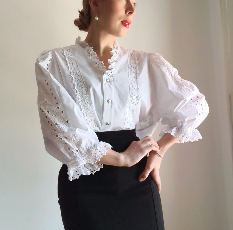 Puede incluir: Blusa blanca de ojal con cuello alto y mangas largas. La blusa presenta intrincados detalles de encaje a lo largo del cuello, la tapeta y las mangas. Las mangas tienen un pu&ntilde;o recogido y con volantes. La blusa se combina con una falda negra.