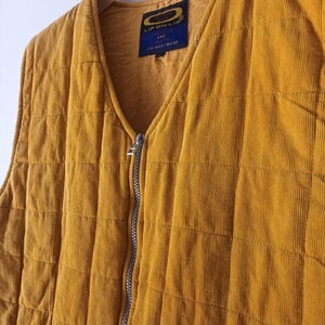May include: Mustard yellow corduroy vest with a quilted design. The vest has a V-neck, a silver zipper, and a label that reads "LIP ON LIP AND FINEST WEAR". The vest is hanging on a white hanger.