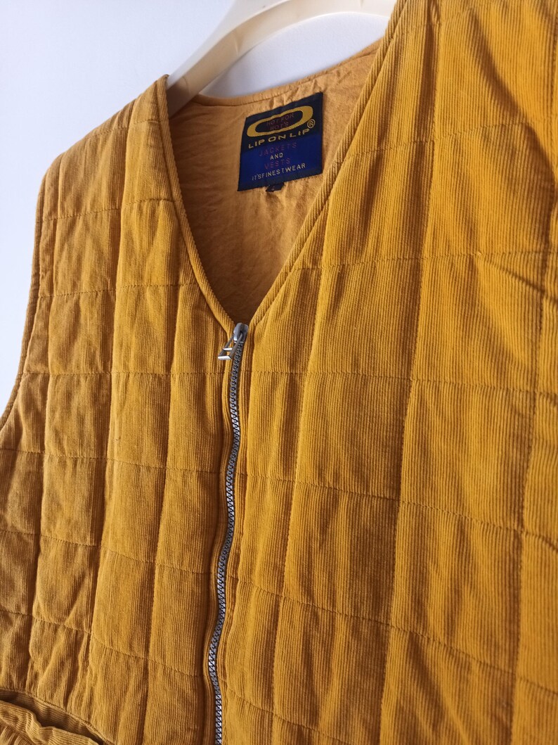 May include: Mustard yellow corduroy vest with a quilted design. The vest has a V-neck, a silver zipper, and a label that reads "LIP ON LIP AND FINEST WEAR". The vest is hanging on a white hanger.