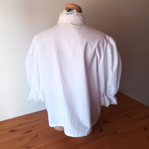 Pode incluir: Uma blusa branca com gola alta e acabamento em renda de ilh&oacute;s intrincada, mangas bufantes volumosas que chegam aos cotovelos e detalhes de renda de ilh&oacute;s a condizer nos punhos. A pe&ccedil;a &eacute; confeccionada em tecido branco s&oacute;lido.