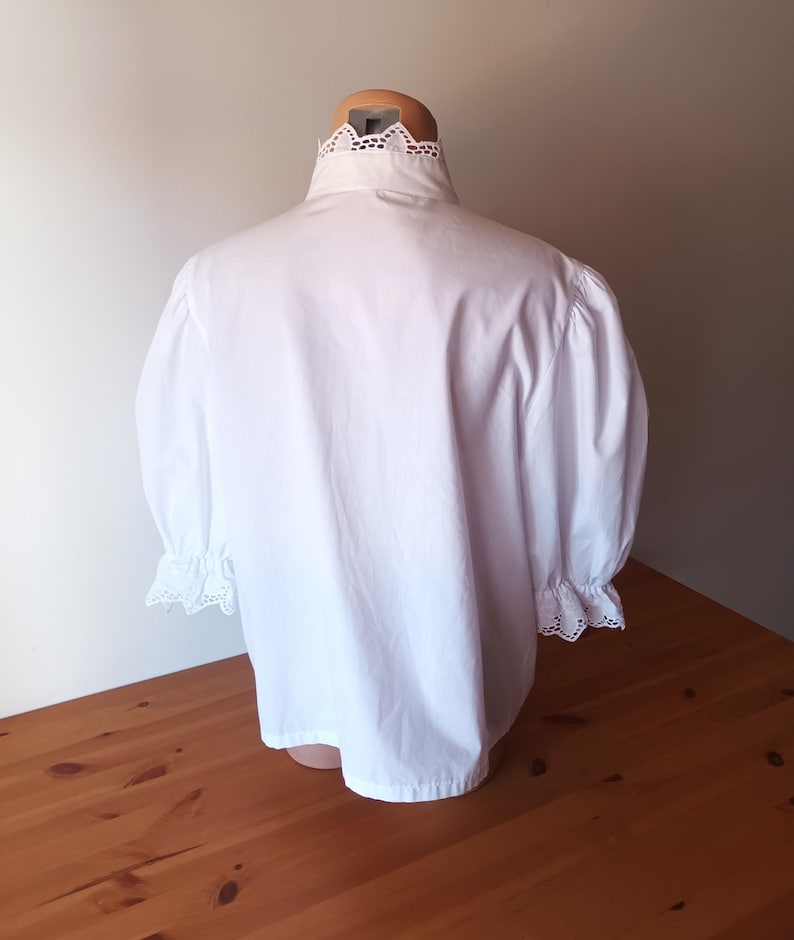 Pode incluir: Uma blusa branca com gola alta e acabamento em renda de ilh&oacute;s intrincada, mangas bufantes volumosas que chegam aos cotovelos e detalhes de renda de ilh&oacute;s a condizer nos punhos. A pe&ccedil;a &eacute; confeccionada em tecido branco s&oacute;lido.