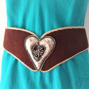 May include: A brown suede belt with a decorative silver heart-shaped buckle featuring a floral design. The belt is accented with a brown rope trim.