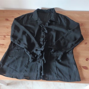 May include: A black sheer blouse with a ruffled neckline and long sleeves. The blouse has a button-up front and a tie at the waist.