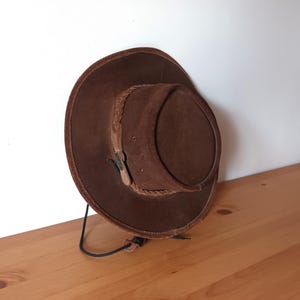 Op de afbeelding: Een bruine suède hoed met een brede rand en een gevlochten band rond de kroon. De hoed heeft een leren riem met een gesp en een kinband. De hoed staat op een houten oppervlak.