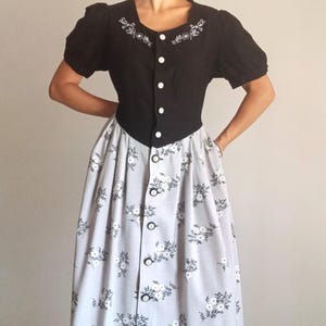 May include: A vintage dress with a black bodice, white buttons, and floral embroidery. The light grey skirt has a white floral pattern. The dress features short, puffed sleeves, offering a classic, retro aesthetic.