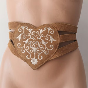 May include: A tan suede belt with a heart-shaped centerpiece. The heart is adorned with white embroidered floral designs and the number "1896". The belt features multiple straps for a secure fit, showcasing a vintage or traditional style.