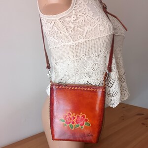 May include: A small brown leather crossbody bag with a floral design. The bag has a zipper closure and a long adjustable strap.