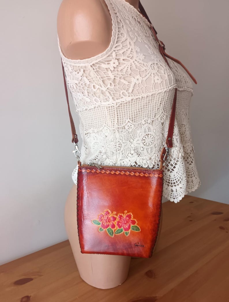May include: A small brown leather crossbody bag with a floral design. The bag has a zipper closure and a long adjustable strap.