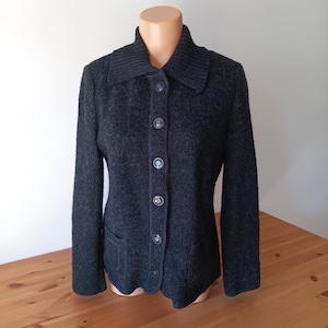 Op de afbeelding: Een donkergrijze wollen jas met een geribbelde kraag en knoopsluiting. De jas heeft twee voorzakken en wordt op een mannequin gepresenteerd. De knopen zijn een lichtere grijstint. Klassieke stijl.