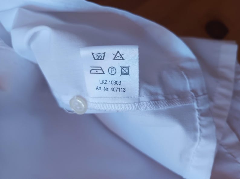 Pode incluir: Uma camisa branca com uma etiqueta de cuidados contendo s&iacute;mbolos de lavagem, incluindo um &iacute;cone de lavagem a 30 graus, um s&iacute;mbolo de n&atilde;o usar lix&iacute;via, um &iacute;cone de ferro, um s&iacute;mbolo de limpeza a seco e um s&iacute;mbolo de secadora. LKZ 10303 e Art.-Nr. 407113.