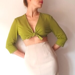Può includere: Un top corto verde chiaro a maniche lunghe con un nodo sul davanti. Il top è abbinato a una gonna beige a vita alta. L'outfit è semplice ed elegante, adatto a un'occasione casual o semi-formale.