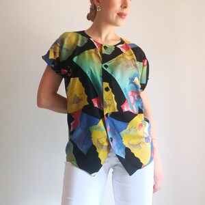 Op de afbeelding: Een shirt met korte mouwen en knoopsluiting, voorzien van een zwarte achtergrond met een levendig, veelkleurig geometrisch patroon in tinten geel, blauw, groen en rood. Het shirt heeft een ronde halslijn en een knoopsluiting aan de voorzijde.