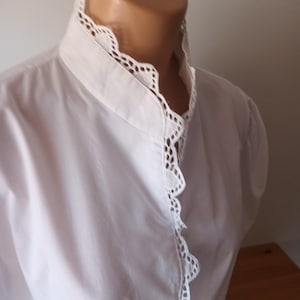 Pode incluir: Uma camisa branca de manga comprida com gola alta e uma carcela frontal adornada com um intrincado acabamento em renda branca festonada. A pe&ccedil;a &eacute; feita de um tecido branco liso e s&oacute;lido, exibindo um ajuste estruturado no pesco&ccedil;o.