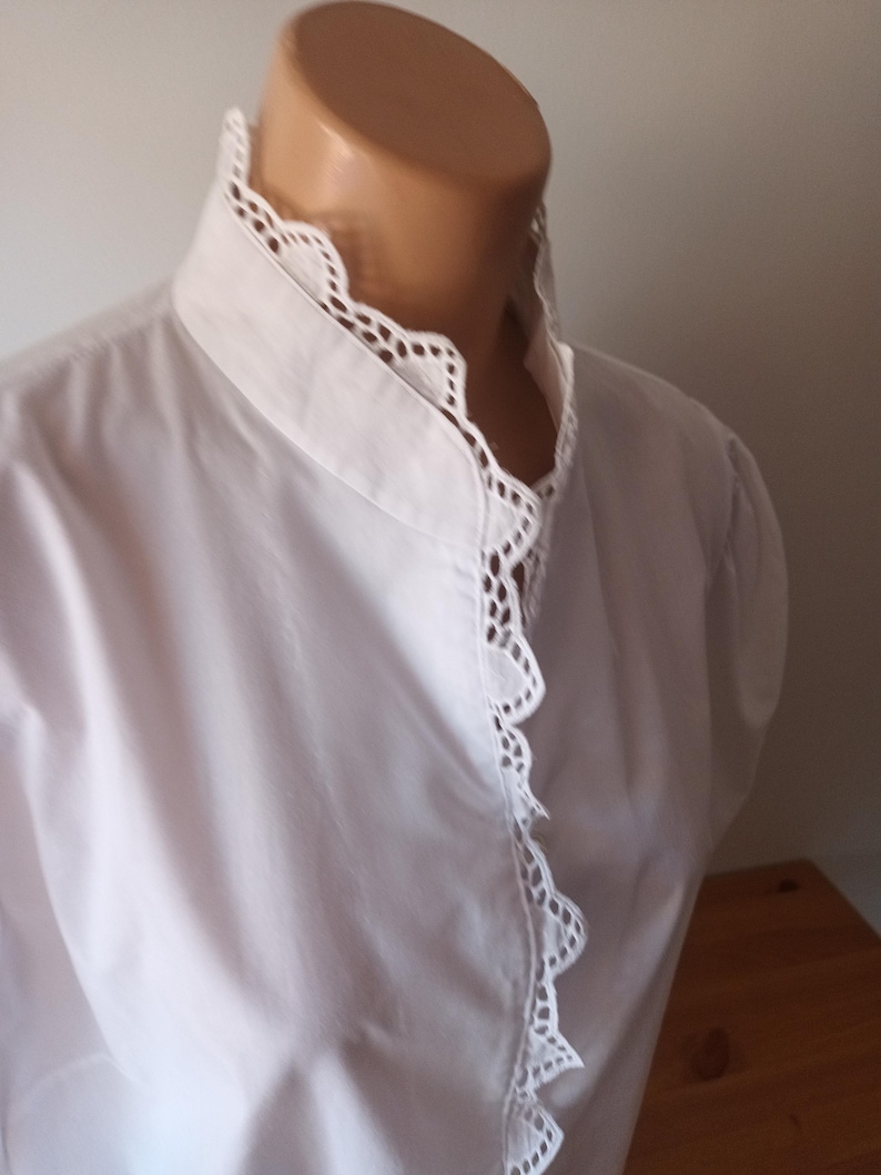 Pode incluir: Uma camisa branca de manga comprida com gola alta e uma carcela frontal adornada com um intrincado acabamento em renda branca festonada. A pe&ccedil;a &eacute; feita de um tecido branco liso e s&oacute;lido, exibindo um ajuste estruturado no pesco&ccedil;o.