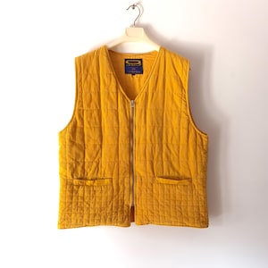 May include: Mustard yellow quilted vest with a V-neck, two front pockets, and a full-length zip. The vest is hanging on a white hanger against a white background. A small label is visible near the neckline.