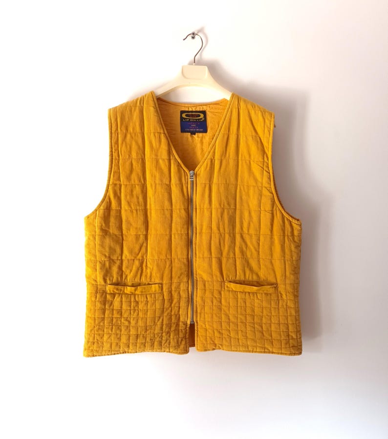 May include: Mustard yellow quilted vest with a V-neck, two front pockets, and a full-length zip. The vest is hanging on a white hanger against a white background. A small label is visible near the neckline.