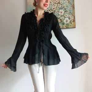 Puede incluir: Blusa negra de manga larga con ribete de volantes y mangas acampanadas. La blusa tiene un escote en V y un corpi&ntilde;o entallado. Las mangas son transparentes y fluidas. La blusa se combina con pantalones blancos.
