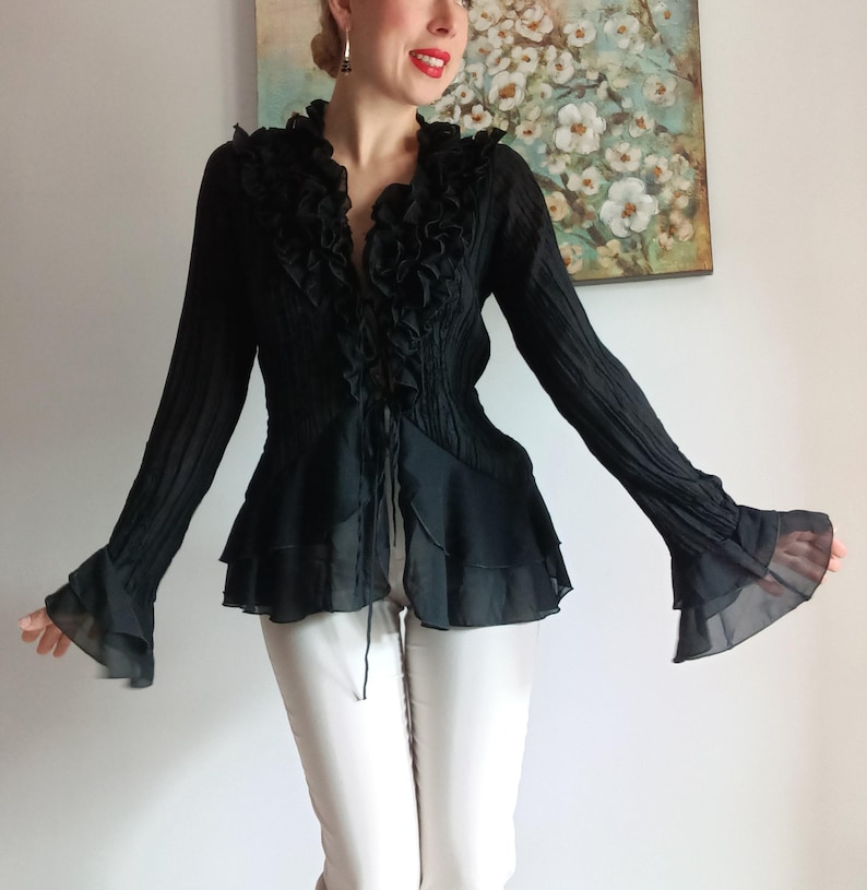 Puede incluir: Blusa negra de manga larga con ribete de volantes y mangas acampanadas. La blusa tiene un escote en V y un corpi&ntilde;o entallado. Las mangas son transparentes y fluidas. La blusa se combina con pantalones blancos.