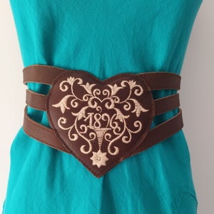 May include: A brown leather belt with a heart-shaped buckle. The buckle is embroidered with a floral design and the year "1896".