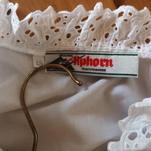 Puede incluir: Blusa blanca con cuello y ribete de encaje calado detallado. La etiqueta dice "Original Alphorn Trachtenmode". Una percha dorada sostiene la blusa. La etiqueta de talla indica "48".