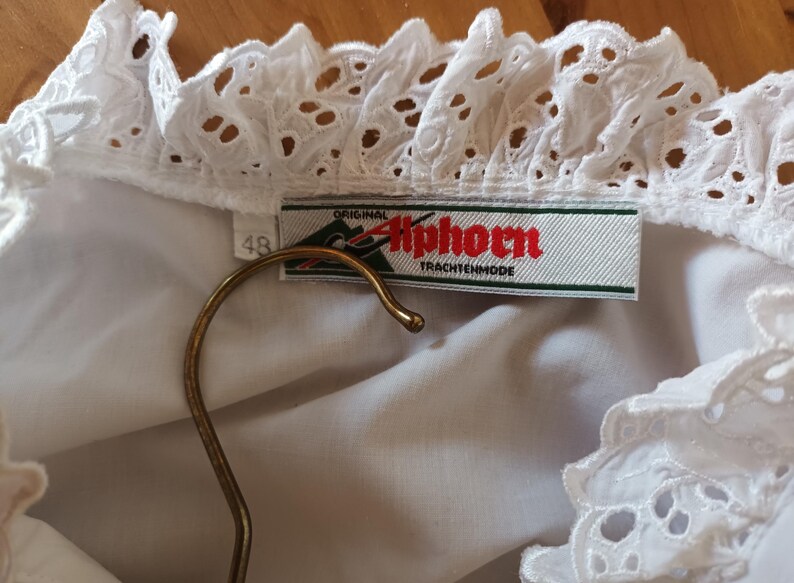 Puede incluir: Blusa blanca con cuello y ribete de encaje calado detallado. La etiqueta dice "Original Alphorn Trachtenmode". Una percha dorada sostiene la blusa. La etiqueta de talla indica "48".