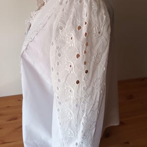 Puede incluir: Blusa blanca con intrincado bordado de ojal en las mangas. La blusa presenta un cuello y pu&ntilde;os con volantes, a&ntilde;adiendo un toque vintage. Las mangas tienen un patr&oacute;n floral con detalles calados.