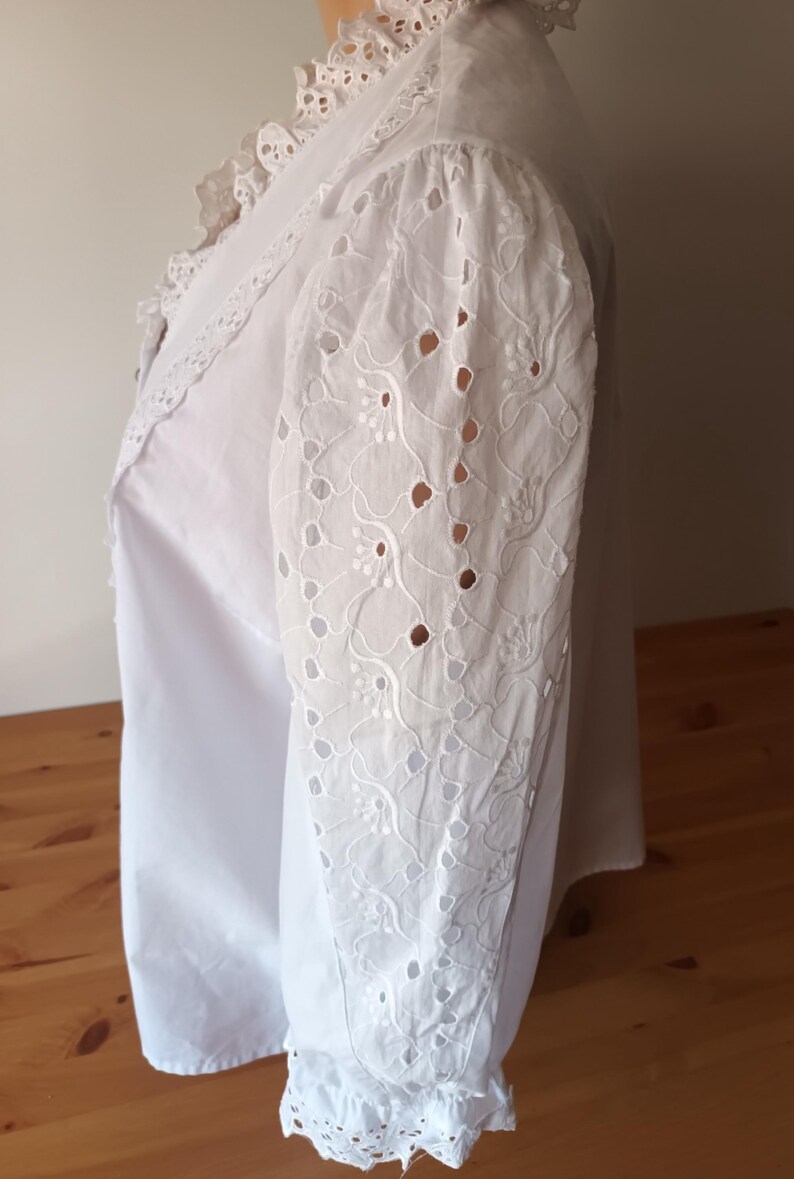 Puede incluir: Blusa blanca con intrincado bordado de ojal en las mangas. La blusa presenta un cuello y pu&ntilde;os con volantes, a&ntilde;adiendo un toque vintage. Las mangas tienen un patr&oacute;n floral con detalles calados.