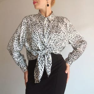 Pode incluir: Camisa estampada em preto e branco amarrada na cintura. A camisa de manga comprida tem gola e é estampada com pequenas formas pretas irregulares sobre um fundo branco. É usada com uma saia preta.