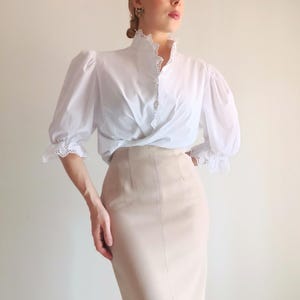 Pode incluir: Uma blusa branca com mangas bufantes, gola alta e acabamento em renda branca ao longo da carcela de bot&otilde;es e punhos, combinada com uma saia l&aacute;pis bege de cintura alta com detalhes de costura vertical na parte frontal.