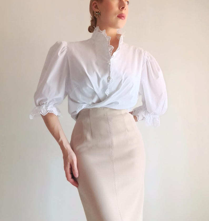 Pode incluir: Uma blusa branca com mangas bufantes, gola alta e acabamento em renda branca ao longo da carcela de bot&otilde;es e punhos, combinada com uma saia l&aacute;pis bege de cintura alta com detalhes de costura vertical na parte frontal.