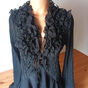 Puede incluir: Blusa negra con cuello con volantes y mangas largas. La blusa presenta un escote en V profundo y un corpi&ntilde;o ajustado con detalles fruncidos. Las mangas tienen un dise&ntilde;o acampanado. La blusa se exhibe en un maniqu&iacute;.