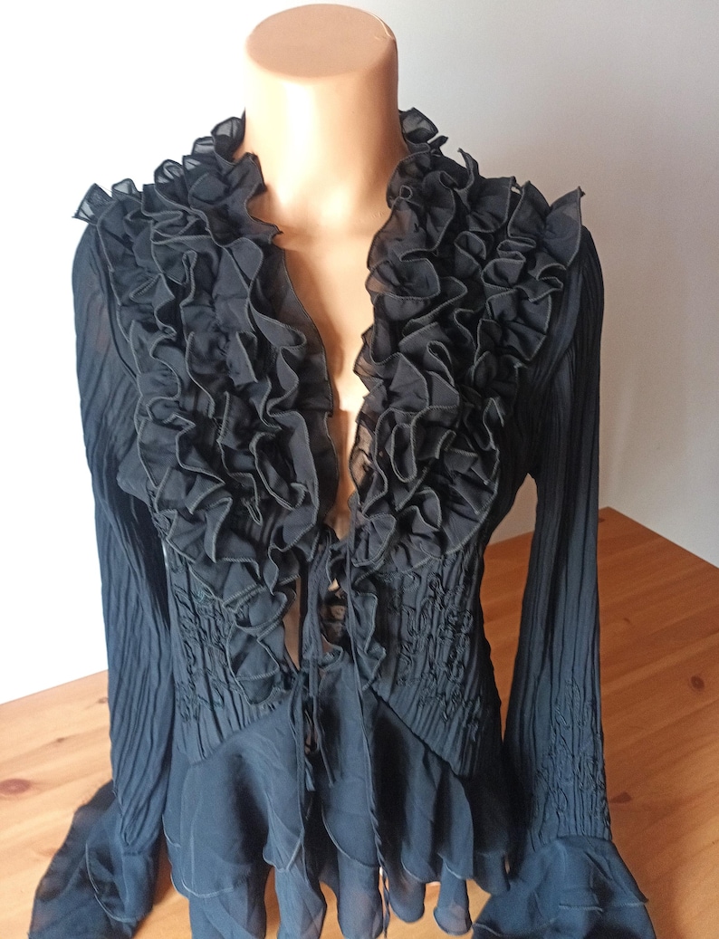 Puede incluir: Blusa negra con cuello con volantes y mangas largas. La blusa presenta un escote en V profundo y un corpi&ntilde;o ajustado con detalles fruncidos. Las mangas tienen un dise&ntilde;o acampanado. La blusa se exhibe en un maniqu&iacute;.