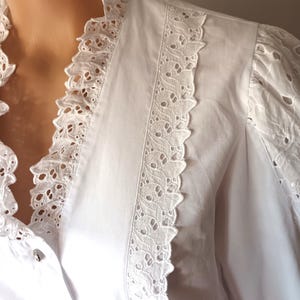 Puede incluir: Una blusa blanca con intrincados detalles de encaje calado. La blusa presenta un cuello con volantes y adornos de encaje en la parte delantera y las mangas. La tela parece ser de algod&oacute;n o lino ligero, con peque&ntilde;os botones decorativos.