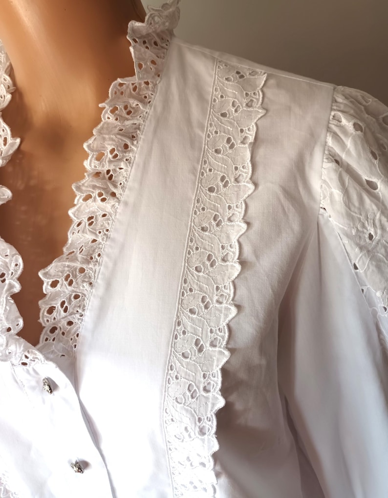 Puede incluir: Una blusa blanca con intrincados detalles de encaje calado. La blusa presenta un cuello con volantes y adornos de encaje en la parte delantera y las mangas. La tela parece ser de algod&oacute;n o lino ligero, con peque&ntilde;os botones decorativos.