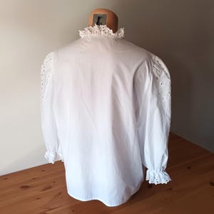 Puede incluir: Una blusa blanca con cuello alto con volantes y mangas largas. Las mangas presentan detalles de ojal y terminan en pu&ntilde;os con volantes. La blusa se exhibe en un maniqu&iacute;, mostrando su dise&ntilde;o cl&aacute;sico y textura.