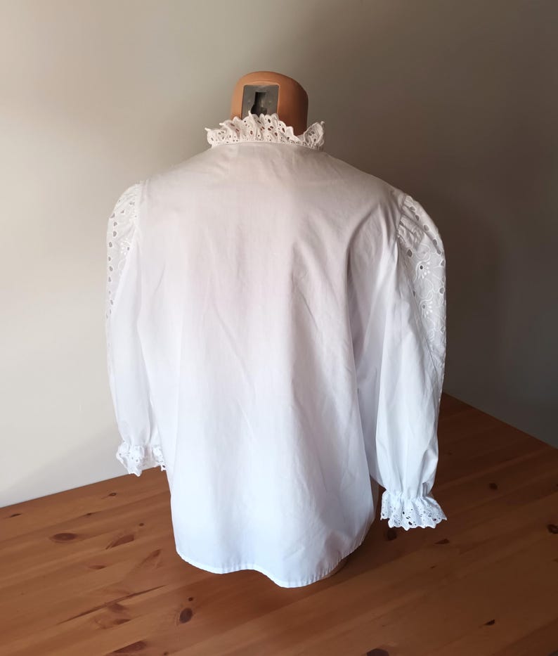 Puede incluir: Una blusa blanca con cuello alto con volantes y mangas largas. Las mangas presentan detalles de ojal y terminan en pu&ntilde;os con volantes. La blusa se exhibe en un maniqu&iacute;, mostrando su dise&ntilde;o cl&aacute;sico y textura.