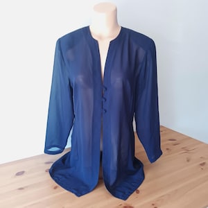 May include: A navy blue, long-sleeved jacket with a collarless design and three buttons down the front. The sheer fabric drapes over a mannequin, showcasing the garment's flowing silhouette. The jacket is displayed on a wooden surface.