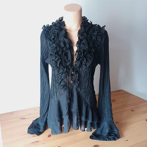 Puede incluir: Blusa negra con cuello y pu&ntilde;os con volantes. La parte superior de manga larga presenta un corpi&ntilde;o plisado y un dise&ntilde;o fluido y en capas. La blusa se muestra en un maniqu&iacute;.