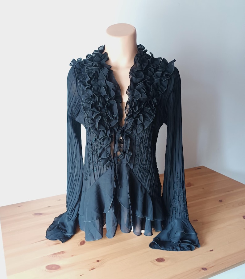 Puede incluir: Blusa negra con cuello y pu&ntilde;os con volantes. La parte superior de manga larga presenta un corpi&ntilde;o plisado y un dise&ntilde;o fluido y en capas. La blusa se muestra en un maniqu&iacute;.