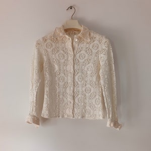 May include: A white crochet blouse with long sleeves and a button-up front. The blouse has a delicate floral pattern and a ruffled trim on the cuffs.