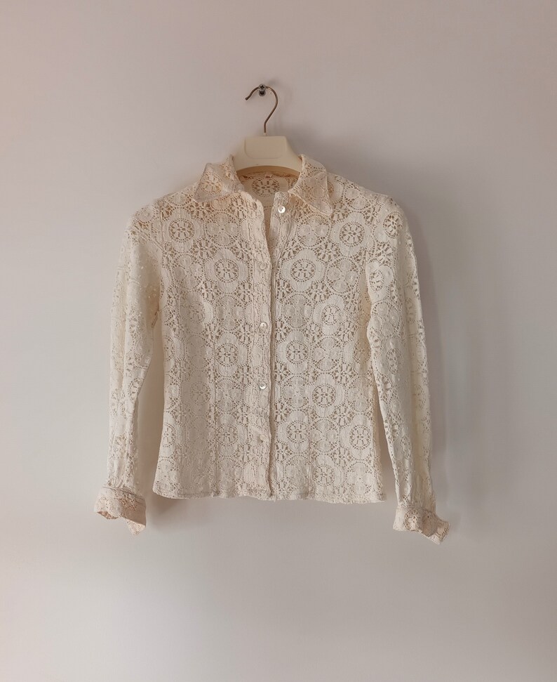 May include: A white crochet blouse with long sleeves and a button-up front. The blouse has a delicate floral pattern and a ruffled trim on the cuffs.