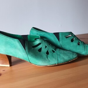 Puede incluir: Un par de botines de ante verde con detalles calados. Los zapatos tienen punta afilada, tacón bajo de madera y paneles laterales elásticos negros. Los botines se exhiben sobre una superficie de madera.