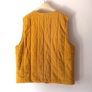 May include: Mustard yellow quilted vest hanging on a white hanger. The vest features a square quilted pattern and a round neckline. The fabric appears to be corduroy. The sleeveless vest has a simple, classic design.