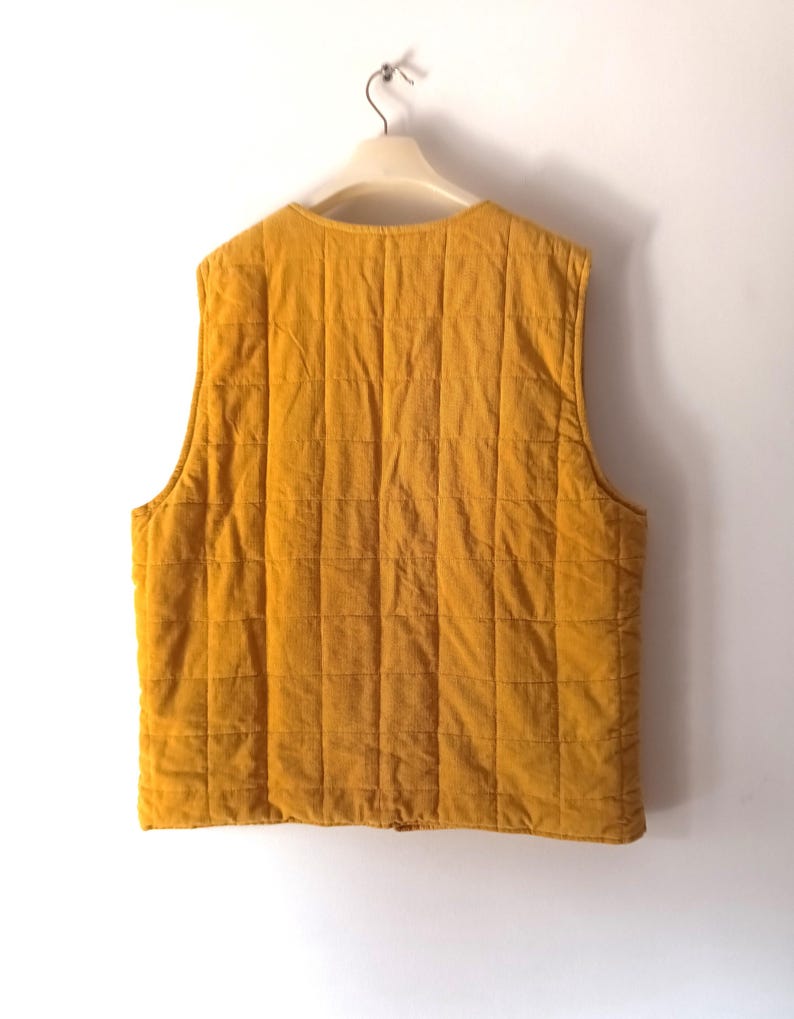 May include: Mustard yellow quilted vest hanging on a white hanger. The vest features a square quilted pattern and a round neckline. The fabric appears to be corduroy. The sleeveless vest has a simple, classic design.