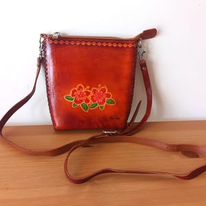 May include: A brown leather crossbody bag with a floral design. The bag has a long adjustable strap and a zipper closure.