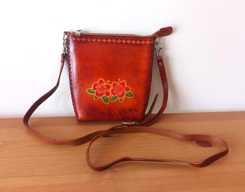 May include: A brown leather crossbody bag with a floral design. The bag has a long adjustable strap and a zipper closure.