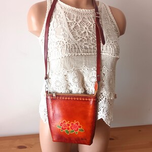 May include: A small, orange leather crossbody bag with a floral design. The bag has a zipper closure and a long adjustable strap.