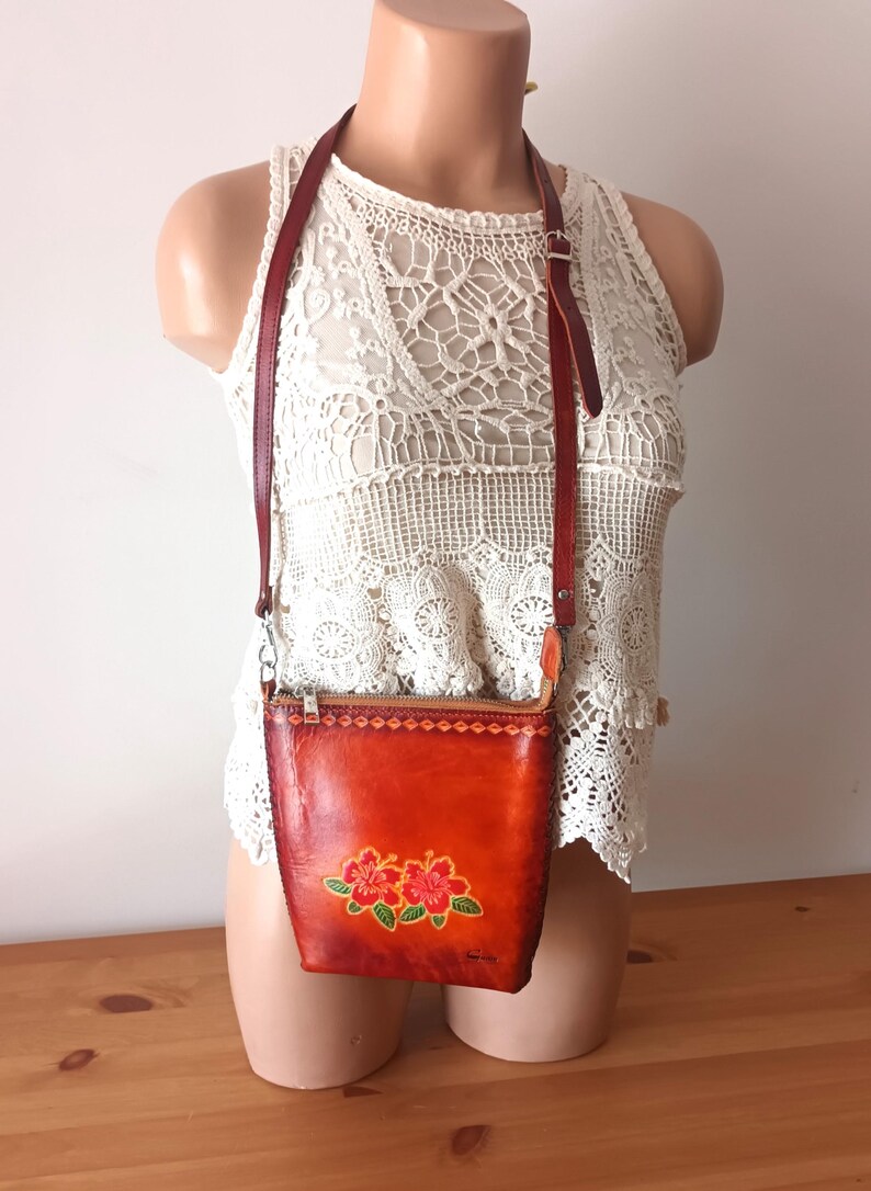 May include: A small, orange leather crossbody bag with a floral design. The bag has a zipper closure and a long adjustable strap.