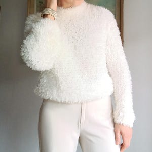 Puede incluir: Un suéter blanco y esponjoso con mangas largas y cuello alto. El suéter se combina con pantalones color crema. Una pulsera con múltiples filas de pequeñas cuentas brillantes es visible en la muñeca izquierda. El atuendo es simple y elegante.