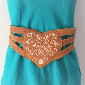 May include: A brown leather belt with a heart-shaped buckle. The buckle is embroidered with a floral design and the year "1896".